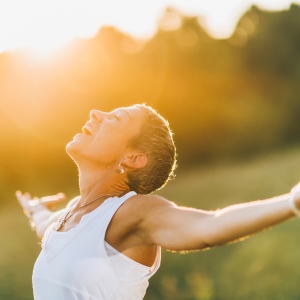 Frau mit ausgebreiteten Armen genießt die Sonne auf einer Wiese – Symbol für neue Energie, Lebensfreude und Gesundheit.