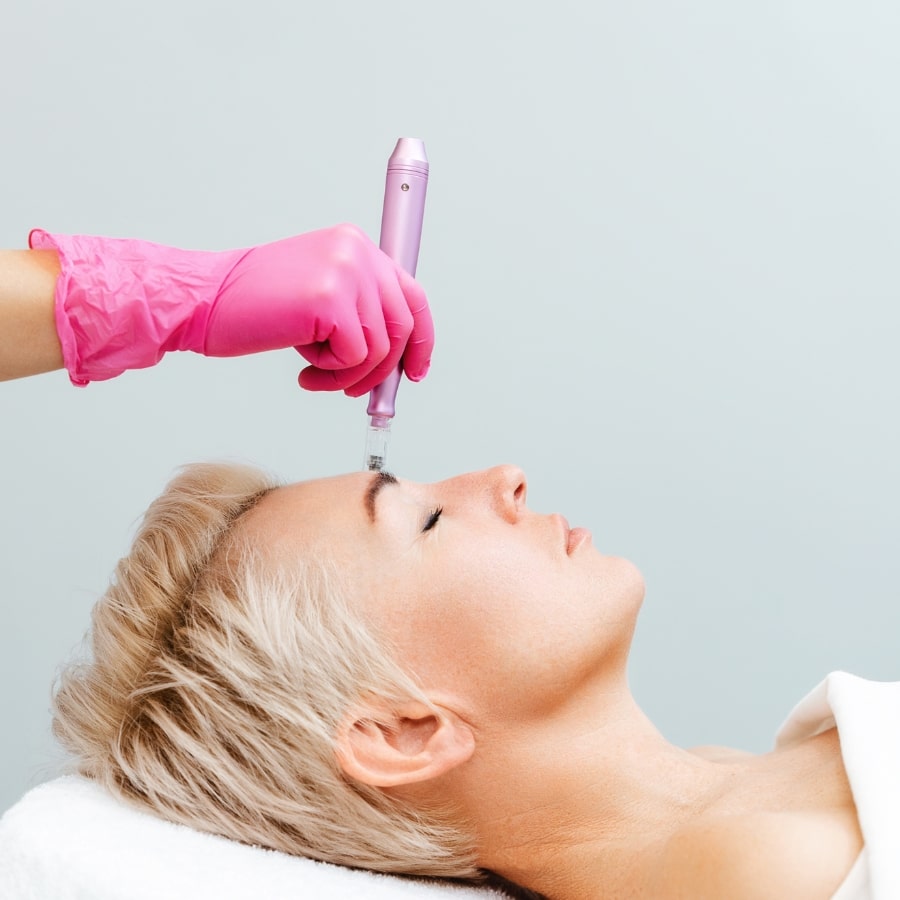 Frau bei einer Stirnbehandlung mit einem ästhetischen Injektionsgerät – Faltenunterspritzung durch Fachpersonal mit pinken Handschuhen.