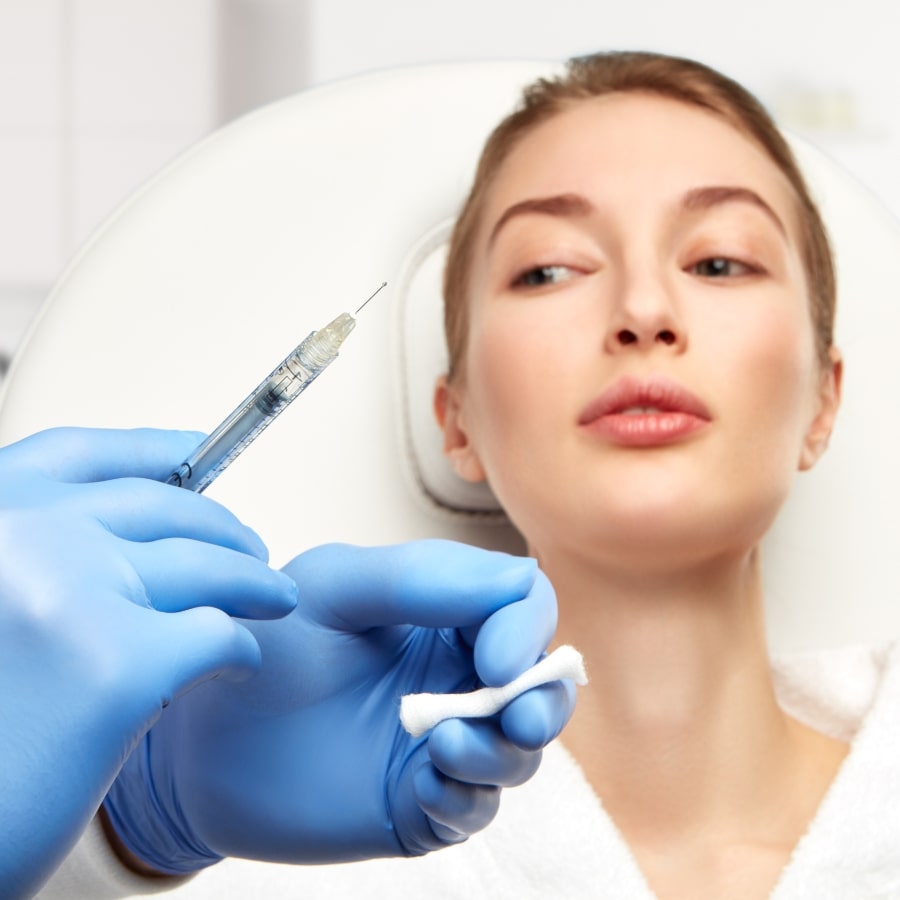 Frau in einer ästhetischen Praxis blickt auf eine vorbereitete Injektionsspritze – Behandlung zur Faltenunterspritzung mit Hyaluronsäure.