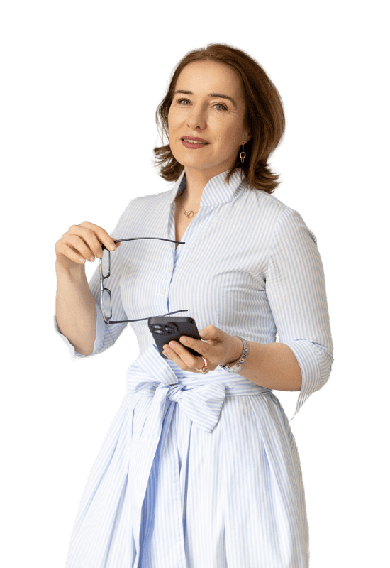 Malgorzata Heinze in einem hellblauen Kleid, mit Brille in der Hand und Smartphone – Ihre Heilpraktikerin mit ganzheitlichem Blick.