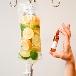 Vitamininfusion mit frischen Zitrusscheiben in einem Infusionsbeutel, daneben eine Hand mit einer Injektionsampulle – Symbol für Vitalstofftherapie und Vitamin-Shots.